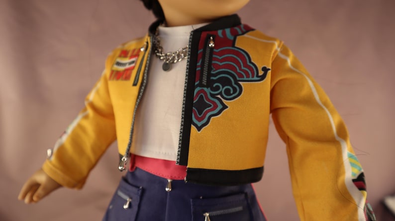 Op de afbeelding: Een pop draagt een gele jas met zwarte afwerking en een decoratief ontwerp aan de voorkant. De jas heeft een ritssluiting en witte strepen op de mouwen. De pop draagt ook een witte top, een zilveren ketting en een blauwe spijkerbroek.