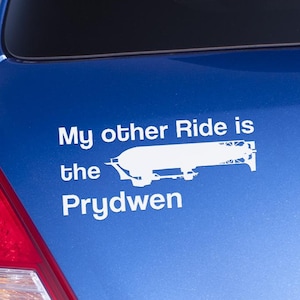 May include: A white vinyl decal on a blue car. The decal reads "My other Ride is the Prydwen" with a stylized image of an airship. The car's taillight is visible on the left.
