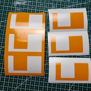 Op de afbeelding: Vier witte stickers met oranje L-vormige symbolen. De stickers zijn bedoeld voor gebruik op voertuigen om een ​​beginnende bestuurder aan te geven.
