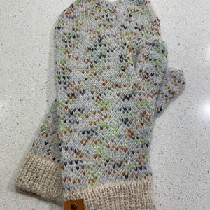May include: Knitted mittens with a speckled pattern in blue, green, brown, and black. The cuffs are cream-coloured, and a small leather tag with the text "Sankta Cecilia" is attached. The mittens are designed for cold weather.