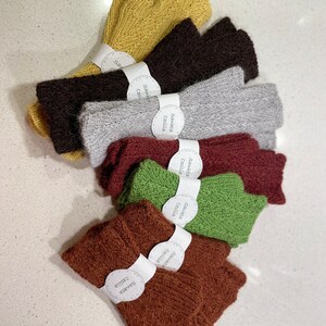 May include: A set of eight knitted fingerless gloves in various colours, including brown, green, red, grey, and yellow. Each glove has a white tag attached with the text "Sawaneta Cecilia" in black.