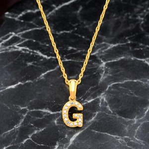 Puede incluir: Collar dorado con un colgante con la forma de la letra "G". El colgante está incrustado con pequeñas piedras preciosas transparentes. La cadena es delicada y tiene un diseño de cuerda trenzada. El fondo es una superficie oscura y veteada.