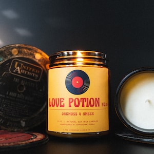 May include: A yellow candle jar with a black and red vinyl record design. The label reads "Love Potion No. 9" and "Darkmoss & Amber". The candle is surrounded by vintage vinyl records.