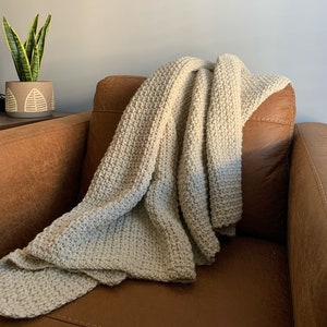 May include: A light gray knitted throw blanket draped over a brown leather armchair.
