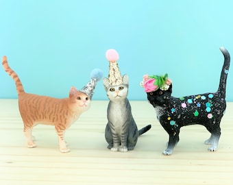 May include: Three cat figurines wearing party hats. One cat is wearing a pink and silver party hat, another is wearing a blue and silver party hat, and the third cat is wearing a flower crown.