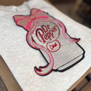 May include: A light gray sweatshirt featuring a sequined Dr Pepper design with a pink bow and the word "Dior". The design is outlined in black and red.