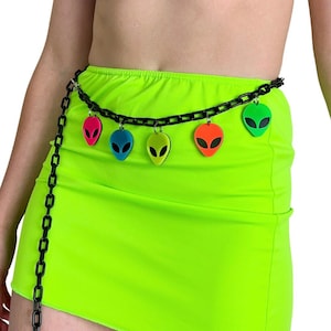 Op de afbeelding: Een zwarte kettingriem met kleurrijke alien-hoofdcharms. De riem wordt gedragen om de taille van een persoon die een neon groene rok draagt.
