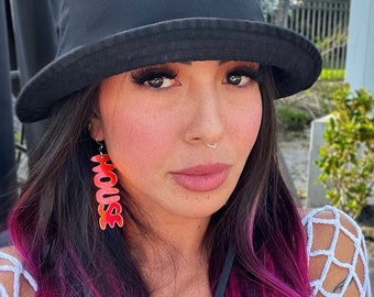 May include: A woman with pink and black hair is wearing a black bucket hat and a white crocheted top. She is wearing large red earrings that spell out "HOUSE".
