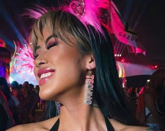 May include: A black and pink techno-themed outfit, including a halter top with chain accents and matching arm sleeves. The outfit features the word "TECHNO" in a bold font. The person is wearing a pink feather headband and clear earrings that say "TECHNO".