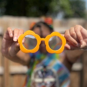 Op de afbeelding: Een paar oranje plastic zonnebrillen in de vorm van een bloem met een gaasontwerp.