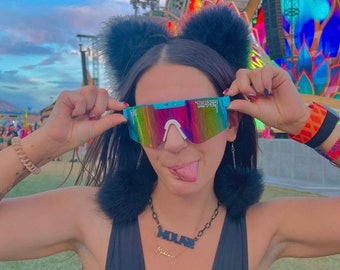May include: A woman wearing a black tank top with a black chain necklace that says "HOUSE" and a gold necklace that says "Mikala". She is wearing iridescent mirrored sunglasses with the word "TRIFECT" printed on the right lens. She is wearing black fuzzy earmuffs with black pom-poms.