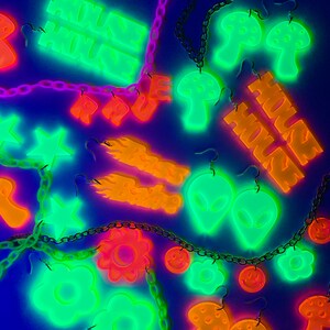May include: A collection of glow-in-the-dark earrings featuring various designs, including mushrooms, stars, flowers, and the words "House House". The earrings are displayed on a black background with chains.