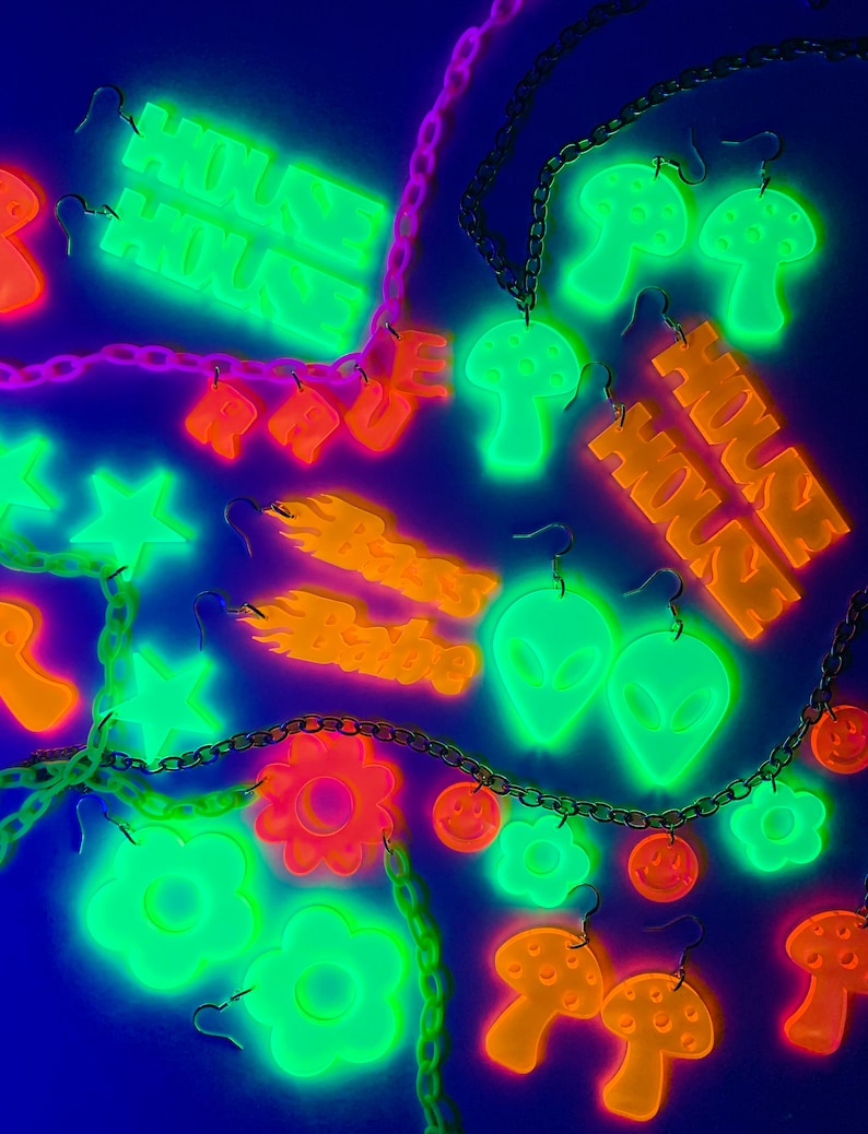 May include: A collection of glow-in-the-dark earrings featuring various designs, including mushrooms, stars, flowers, and the words "House House". The earrings are displayed on a black background with chains.