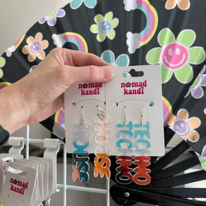 May include: Two pairs of colorful, iridescent earrings with the words "LOVE" and "TECHNO" on them. The earrings are on a white card with the words "Nomad Kandi" printed on them.