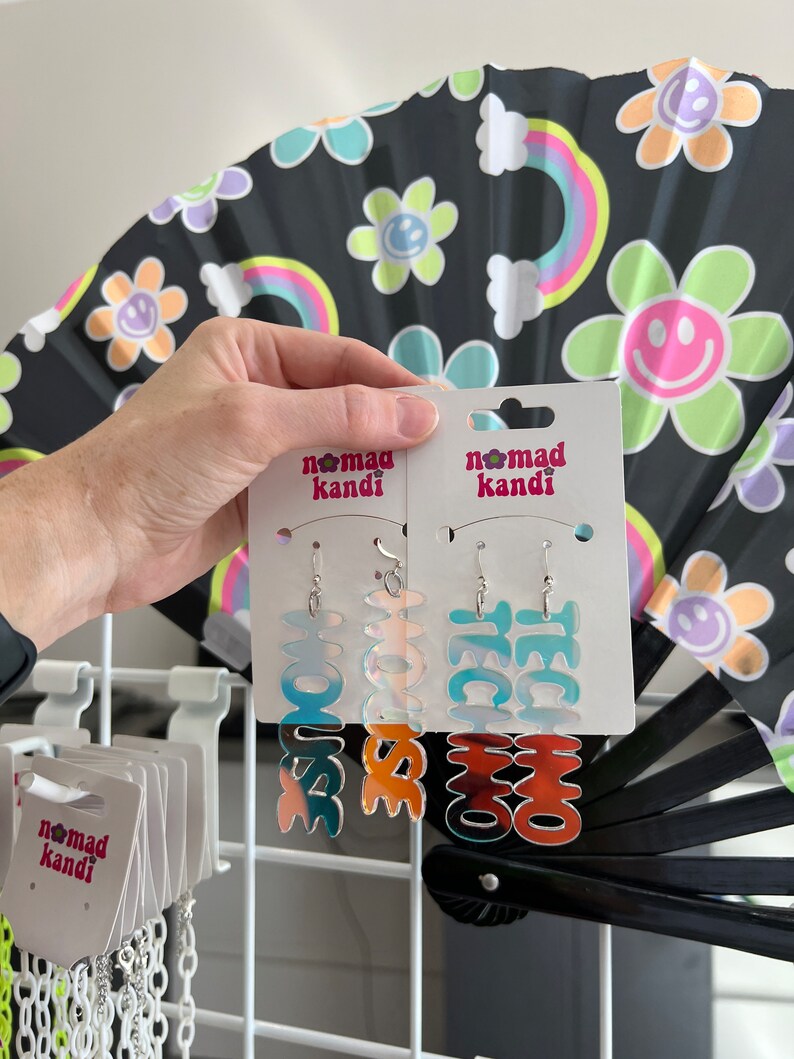 May include: Two pairs of colorful, iridescent earrings with the words "LOVE" and "TECHNO" on them. The earrings are on a white card with the words "Nomad Kandi" printed on them.