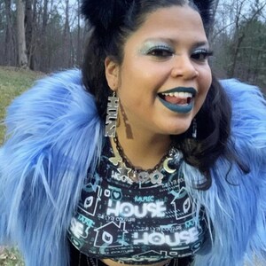 May include: A person wearing a blue faux fur jacket with black fuzzy earmuffs. They are wearing a black crop top with a graphic print that says "House" and a silver chain belt. They have blue eyeshadow and dark blue lipstick.