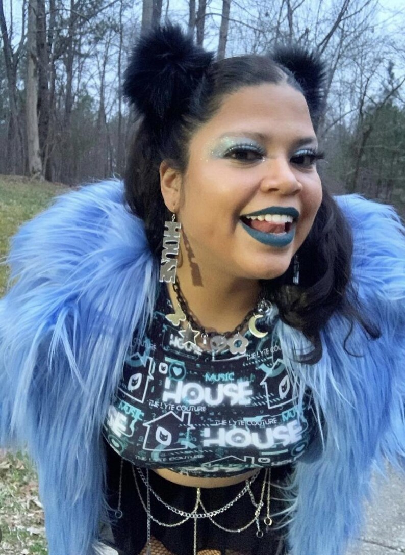 May include: A person wearing a blue faux fur jacket with black fuzzy earmuffs. They are wearing a black crop top with a graphic print that says "House" and a silver chain belt. They have blue eyeshadow and dark blue lipstick.