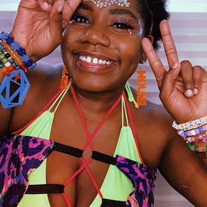 May include: A person wearing a neon green and black halter top with red straps and a purple and black patterned jacket. They are wearing colourful beaded bracelets and earrings that say "HOUSE".