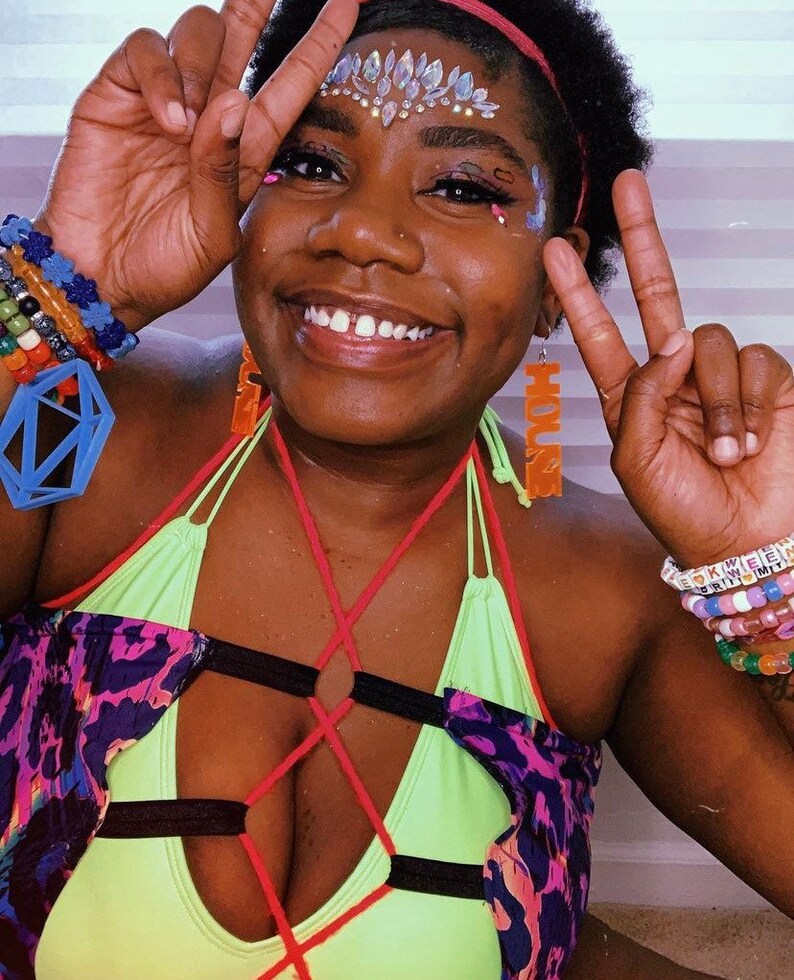 May include: A person wearing a neon green and black halter top with red straps and a purple and black patterned jacket. They are wearing colourful beaded bracelets and earrings that say "HOUSE".