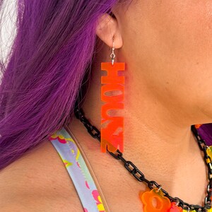 May include: Orange acrylic earrings with the word "HOUSE" spelled out in large letters.