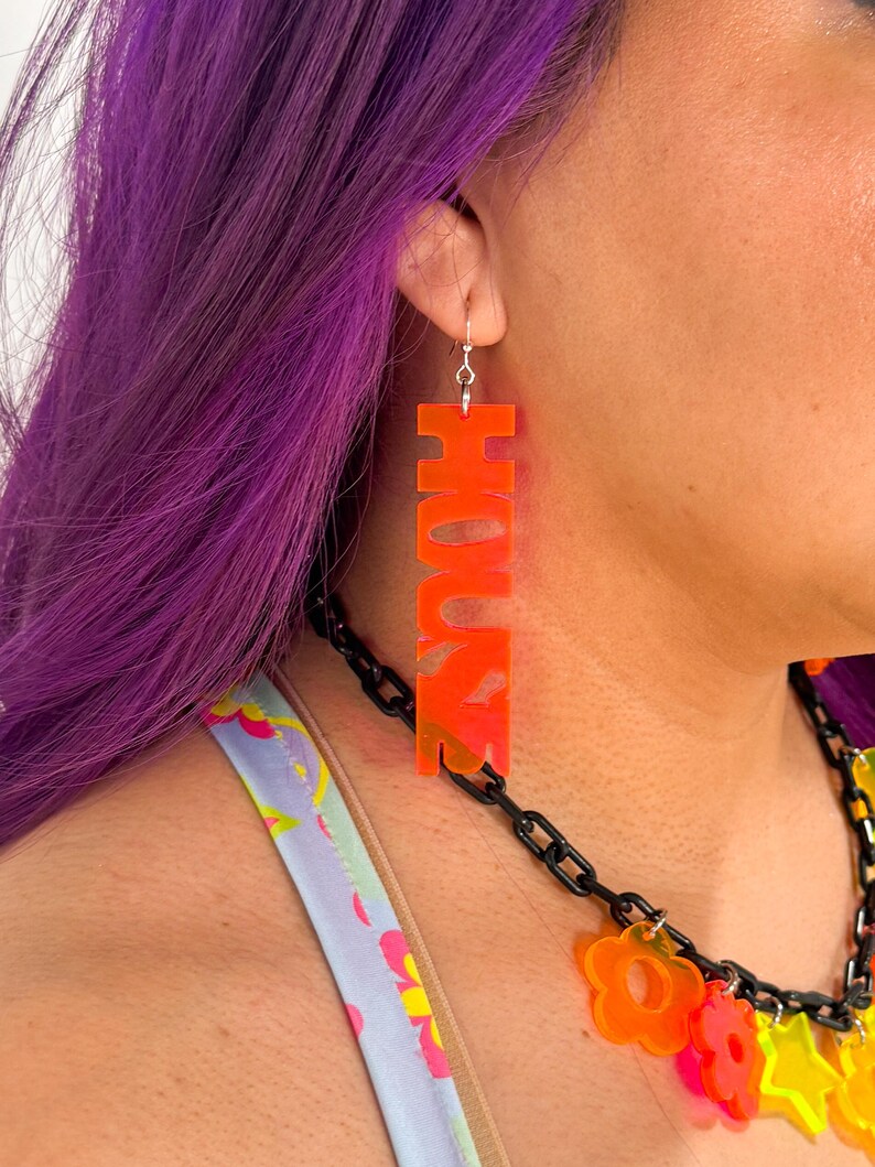 May include: Orange acrylic earrings with the word "HOUSE" spelled out in large letters.