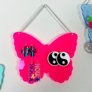 May include: Pink butterfly-shaped earring holder with a silver chain for hanging. The holder has three pairs of earrings displayed: mushroom-shaped earrings, yin-yang earrings, and iridescent earrings with the word "HOUSE" on them.