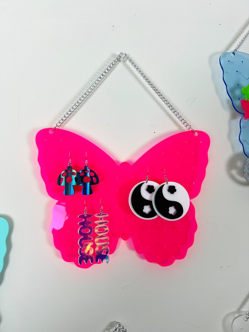 May include: Pink butterfly-shaped earring holder with a silver chain for hanging. The holder has three pairs of earrings displayed: mushroom-shaped earrings, yin-yang earrings, and iridescent earrings with the word "HOUSE" on them.