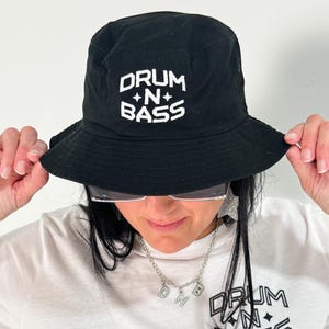 Könnte beinhalten: Schwarzer Fischerhut mit dem weißen Text "DRUM + N + BASS". Der Hut wird von einer Person hochgehalten. Ein weißes T-Shirt mit demselben Text ist ebenfalls sichtbar.