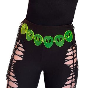 May include: A neon green alien head belt with a chain closure. The belt is worn over black leggings with cut-out detailing. The alien heads are evenly spaced and have a glowing appearance.