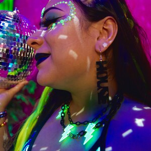 May include: A person with green and black makeup and neon green hair is wearing a black and blue patterned top and a black chain necklace with neon green lightning bolt charms. They are also wearing a long black earring with the word "HOUSE" written on it.