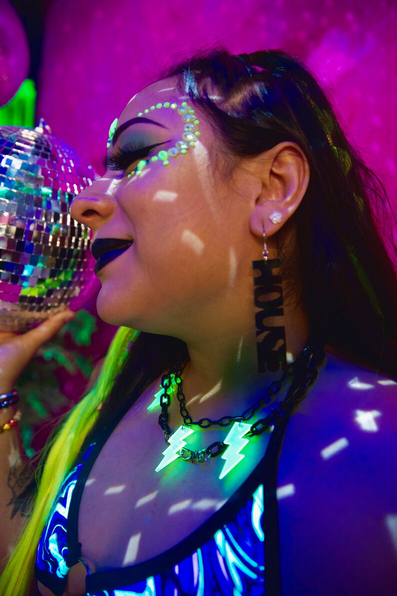 May include: A person with green and black makeup and neon green hair is wearing a black and blue patterned top and a black chain necklace with neon green lightning bolt charms. They are also wearing a long black earring with the word "HOUSE" written on it.