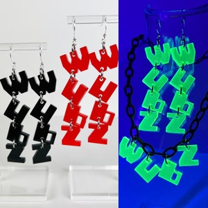 May include: A set of three pairs of earrings featuring the letters "WUZ" in black, red, and glow-in-the-dark green. The earrings are made of acrylic and are attached to silver hooks. The earrings are shown against a white background.