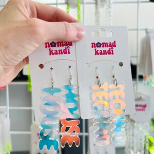 May include: Two pairs of iridescent holographic earrings with the words "House" and "Techno" on them. The earrings are on a white card with the words "Nomad Kande" printed on it.