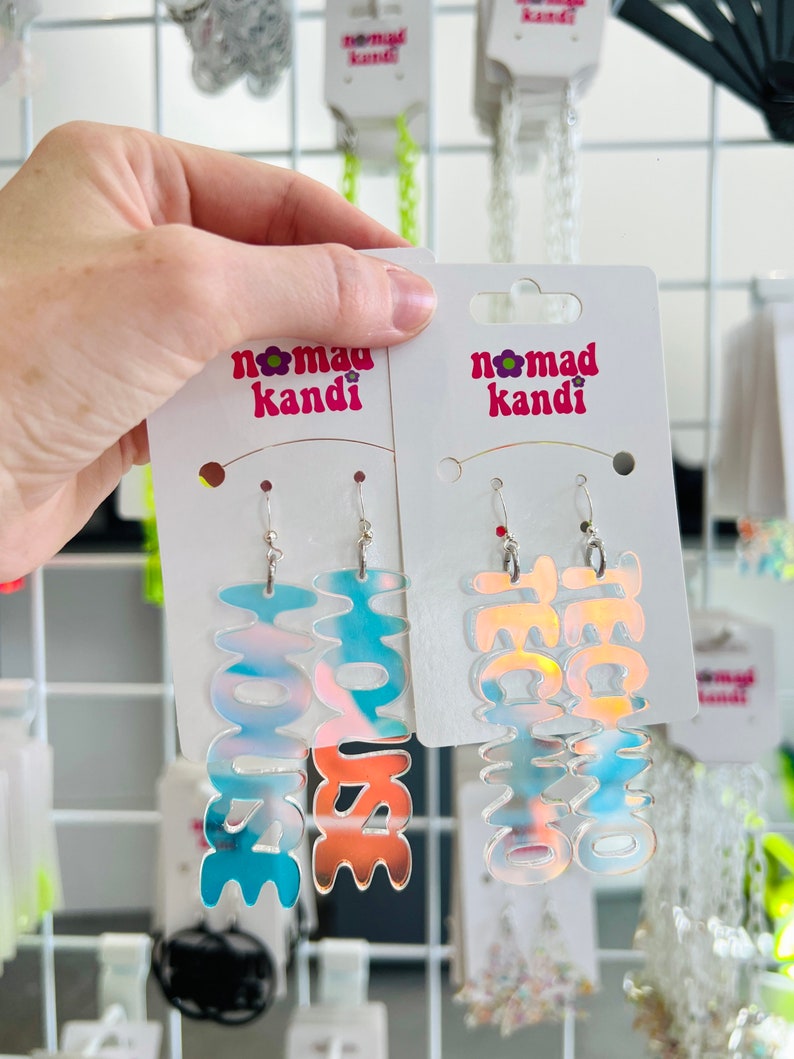 May include: Two pairs of iridescent holographic earrings with the words "House" and "Techno" on them. The earrings are on a white card with the words "Nomad Kande" printed on it.
