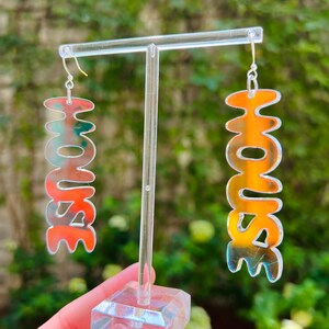 May include: A pair of iridescent silver earrings shaped like the word "HOUSE". The earrings are hanging from a clear plastic earring stand.