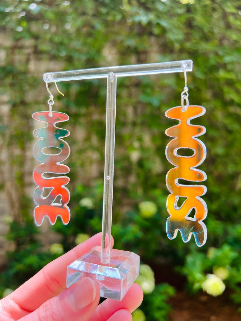 May include: A pair of iridescent silver earrings shaped like the word "HOUSE". The earrings are hanging from a clear plastic earring stand.