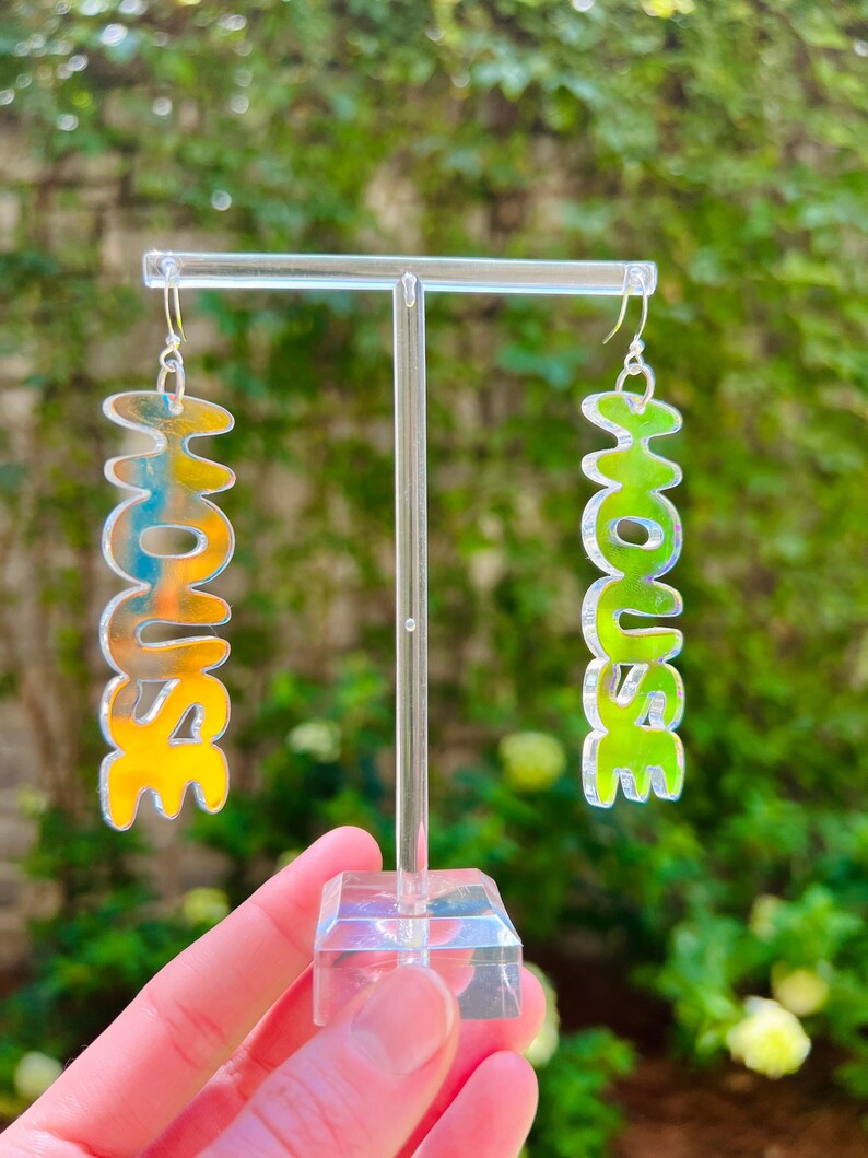 May include: A pair of iridescent earrings with the word "HOUSE" spelled out in a bubbly font. The earrings are displayed on a clear acrylic stand. One earring is yellow and the other is green.
