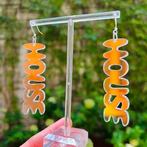 May include: A pair of orange iridescent acrylic earrings with the word "HOUSE" spelled out in a wavy font.