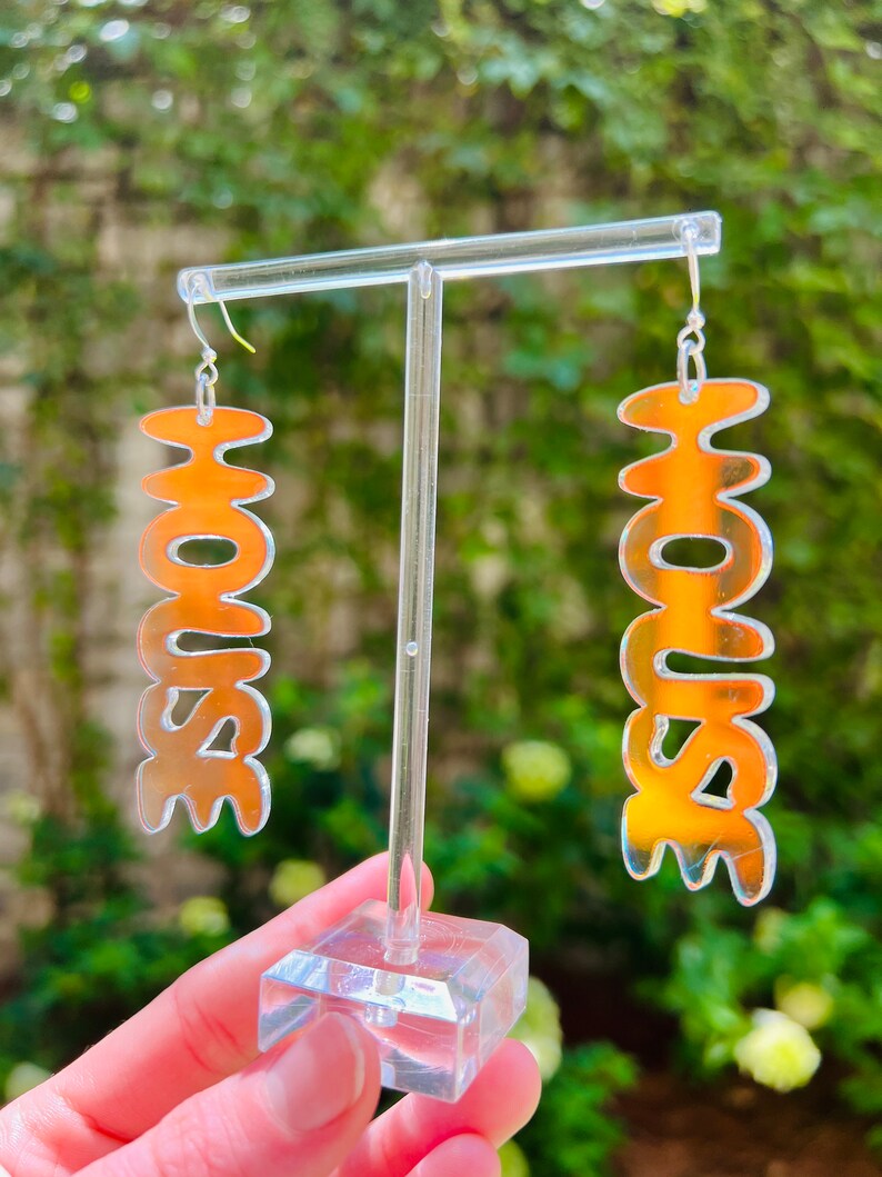 May include: A pair of orange iridescent acrylic earrings with the word "HOUSE" spelled out in a wavy font.
