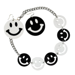 Puede incluir: Un collar de cadena plateada con dijes de caritas sonrientes en blanco y negro. El collar tiene aretes a juego con caritas sonrientes grandes en blanco y negro.