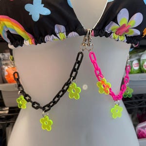 Puede incluir: Un collar de cadena negro con dijes de flores de color verde neón y rosa. La cadena está unida a un top de bikini con estampado floral.