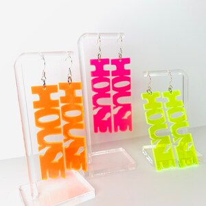 May include: Three pairs of neon acrylic earrings with the word "HOUSE" spelled out in bold letters. The earrings are displayed on clear acrylic stands. The earrings are in pink, orange, and yellow.