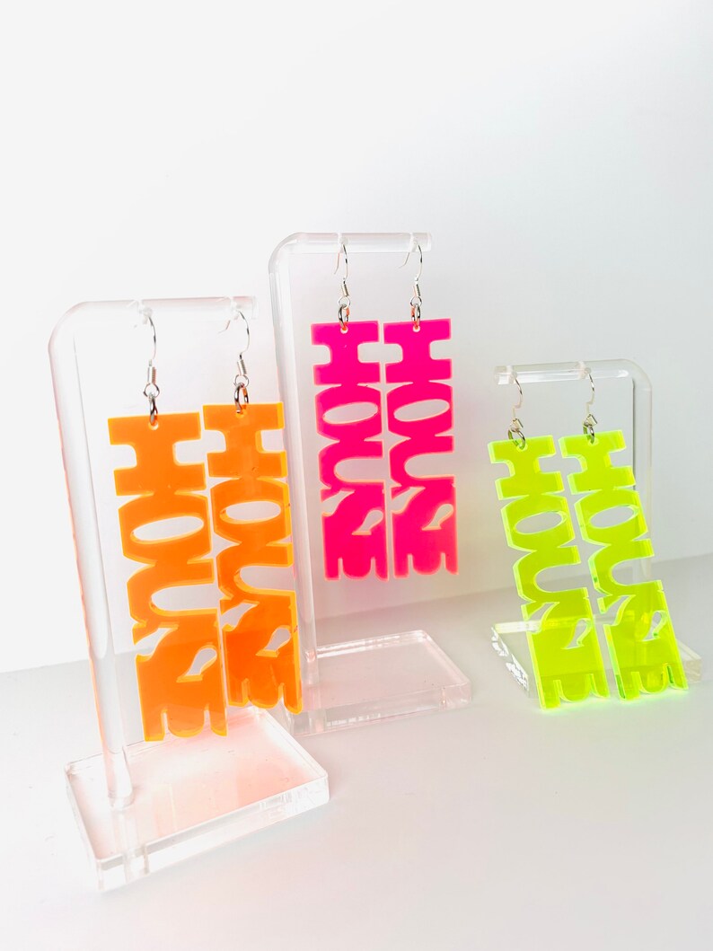 May include: Three pairs of neon acrylic earrings with the word "HOUSE" spelled out in bold letters. The earrings are displayed on clear acrylic stands. The earrings are in pink, orange, and yellow.