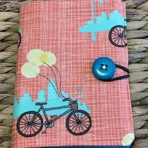 May include: A fabric-covered journal with a pink grid pattern and a blue bicycle design. The journal features a blue button closure and a blue border. The bicycle has a basket and is attached to yellow balloons.