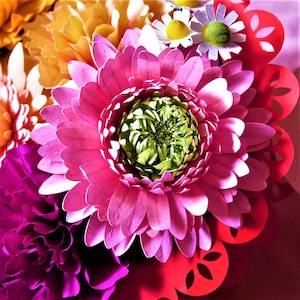 May include: A close-up of a pink paper flower with a green center. The flower is surrounded by other paper flowers in shades of orange, yellow, purple, and red.
