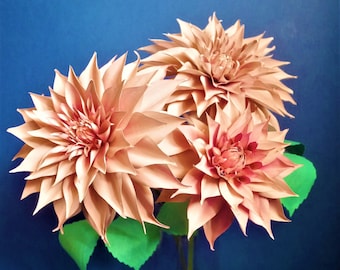 May include: Three pink paper dahlias in a clear glass vase on a blue background. The dahlias have green paper leaves.