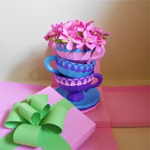 Puede incluir: Una pila de tres tazas de té de papel decoradas con encaje y motivos florales. La taza superior está llena de flores de papel rosa. Las tazas de té están sobre una caja de regalo rosa y verde con un lazo verde.