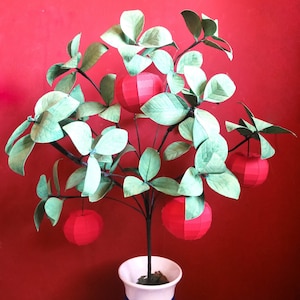 May include: A papercraft tree with green leaves and five red paper apples. The tree is in a white pot.