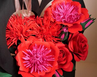 May include: A black heart-shaped box filled with a bouquet of red and pink paper flowers. The flowers are arranged in a variety of shapes and sizes, including roses and peonies.