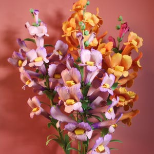 May include: A bouquet of artificial flowers in shades of orange, yellow, and purple. The flowers have a detailed, realistic appearance with green stems and leaves. The background is a solid, muted red.